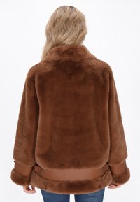 Blonde vrouw gezien van achteren, gekleed in een bruine nep bontjas met leren afwerking en blauwe spijkerbroek tegen een witte achtergrond.