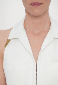 White sleeveless garment with a collar, featuring two front pockets and a visible zipper accentuated by gold hardware details.