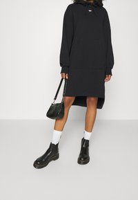 Svart huvtröja klänning med framficka och ribbade ärmslut, parat med svarta ankelstövlar och en liten svart handväska.