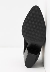 Zapato de tacón alto de ante negro con punta afilada y suela de goma texturizada con un patrón hexagonal. El tacón tiene un diseño de bloque.