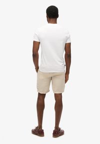 Camiseta blanca de manga corta, pantalones cortos beige con una textura suave. Calzado casual con accents metálicos. Detalle visible en la cinturilla de los pantalones cortos.