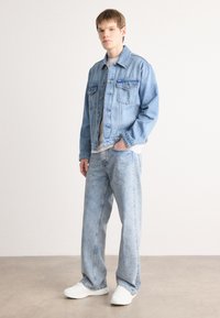 Light-wash denim jacket with two front pockets, worn over a white shirt, paired with matching light-wash wide-leg jeans and white sneakers.