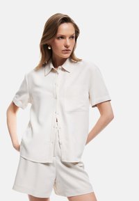 Kortärmad, ljusbeige blus med knäppning framtill, mjuk textur, en br�östficka och avslappnad passform, bärs med matchande shorts.