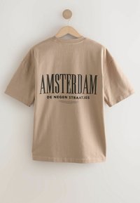 Camiseta de algodón beige colgada en una percha de madera. La parte posterior presenta un texto negro en negrita: "AMSTERDAM" y un texto más pequeño debajo. Mangas cortas, corte relajado.