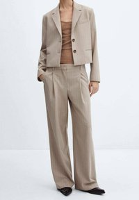 Costume beige à fines rayures comprenant un blazer court à deux boutons avec une poche poitrine, associé à un pantalon  à coupe décontractée, porté sur un haut marron.