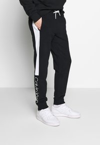 Muž v černých teplácích s bílými bočními pruhy a bílým logem Calvin Klein poblíž kotníku, v bílé obuvi, stojí s rukama v kapsách.