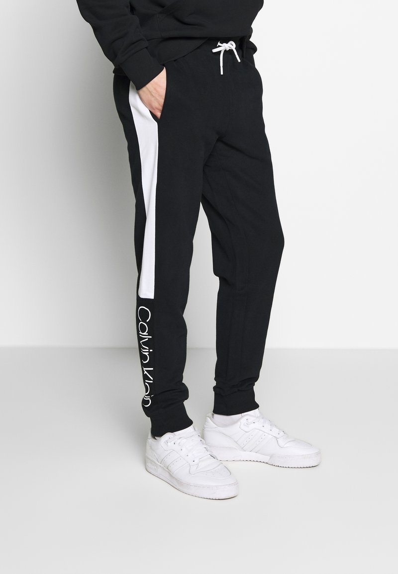 Muž v černých teplácích s bílými bočními pruhy a bílým logem Calvin Klein poblíž kotníku, v bílé obuvi, stojí s rukama v kapsách.