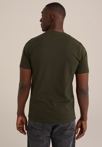 Camiseta de algodón verde oscuro con mangas cortas y escote redondo, combinada con jeans oscuros, vista desde la parte de atrás. Diseño simple, sin patrones visibles.
