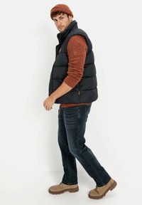 Schwarze Steppweste mit hohem Kragen, getragen über einem orangefarbenen Pullover. Dunkle Jeans und beige Wanderschuhe vervollständigen das Outfit. Eine braune Strickmütze setzt ein zusätzliches Detail.