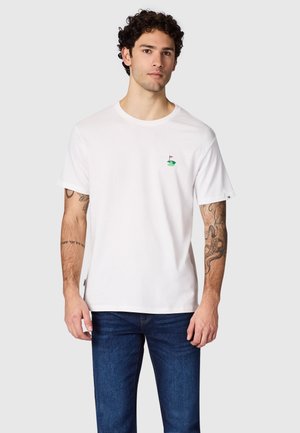 Man met krullend haar draagt een wit T-shirt met een klein groen golfbaanlogo en blauwe spijkerbroek, staand tegen een grijze achtergrond.