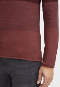 Red knitted sweater featuring a textured pattern, ribbed cuffs, and a leather logo patch at the hem. Worn over dark jeans.