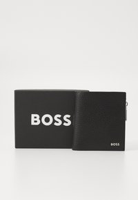 Juodas odinis piniginė su tekstūruotu paviršiumi, turinti sidabrinį užtrauktuką ir iškilnią "BOSS" logotipą, padėta šalia juodos dėžutės su balta užrašyta tekstu.