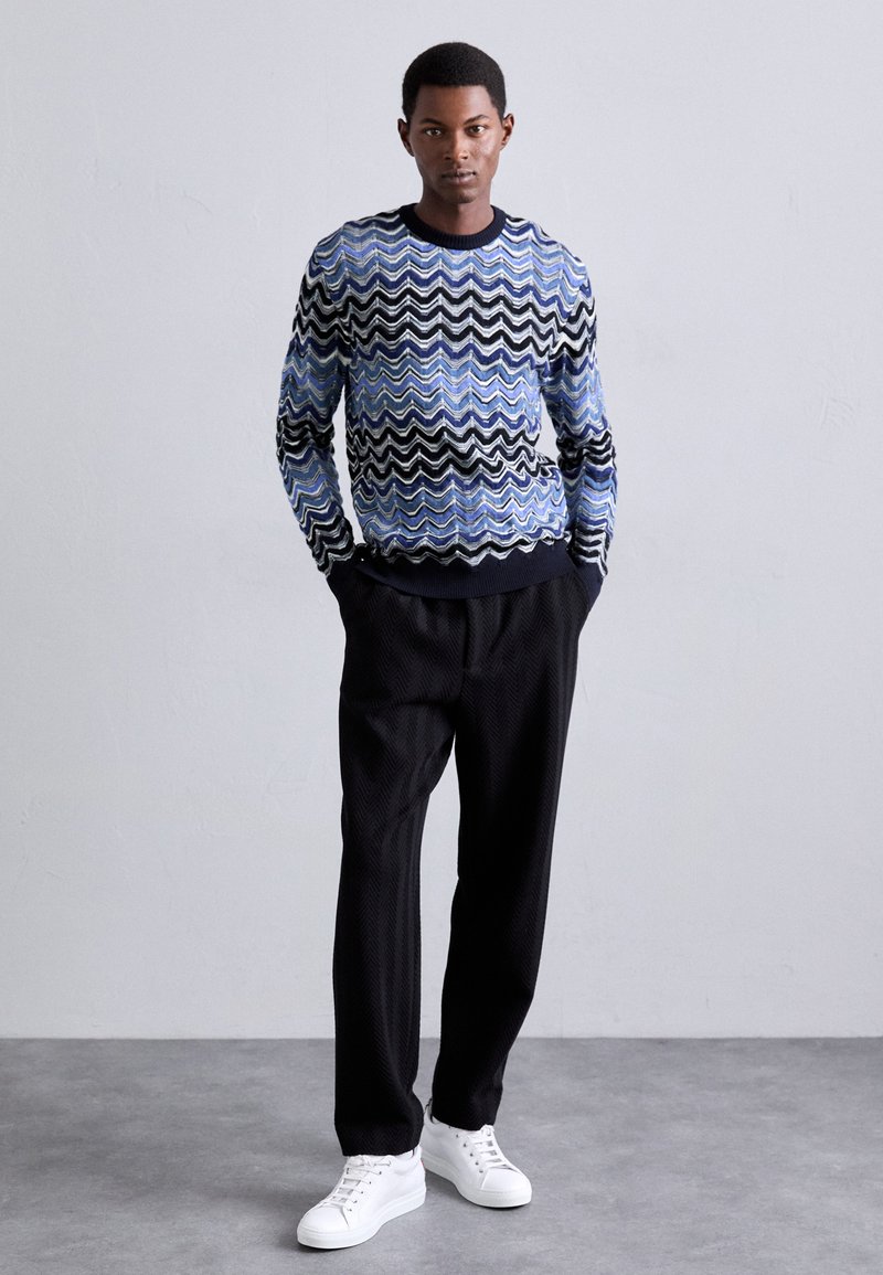 Golf met golven patroon in blauw, zwart en wit. Gecombineerd met gestructureerde zwarte broek en witte sneakers. Model staat tegen een neutrale achtergrond.