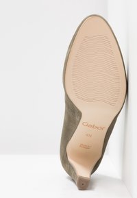 Chaussure en daim vert olive avec un bout pointu, un talon haut et fin, et une semelle en caoutchouc texturée. Caractérisée par un logo de marque embossé et des informations sur la taille.