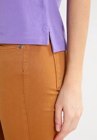 Fialový texturovaný top se bočními rozparky, spárovaný s hnědými přiléhavými kalhotami s viditelným knoflíkem a detaily šití.