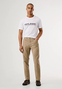 Lichtbeige corduroy broek met een rechte pasvorm, gedragen met een wit T-shirt met kleurrijke merktekst. Bruine veterlaarzen maken de outfit compleet.