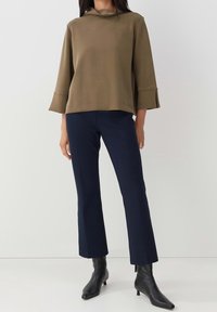 Olivgrön långärmad topp med hög krage och utsvängda ärmar, tillsammans med marinblå byxor och svarta spetsiga klackskor. Slät textur.