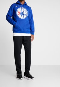 Sweat à capuche Nike bleu avec le logo des 76ers, poche kangourou et cordon de serrage ajustable. Associé à un pantalon noir et des chaussures de sport noires.
