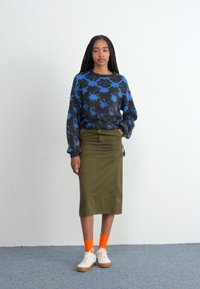 Pleteni pulover z modrim cvetličnim vzorcem, olivno zeleno krilo v obliki svinčnika, oranžne nogavice in bele superge. Model stoji pred nevtralno ozadje.