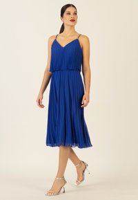 Ein ärmelloses, plissiertes Midi-Kleid in Königsblau mit einem V-Ausschnitt und zarten Trägern, kombiniert mit silbernen, hochhackigen Sandalen.