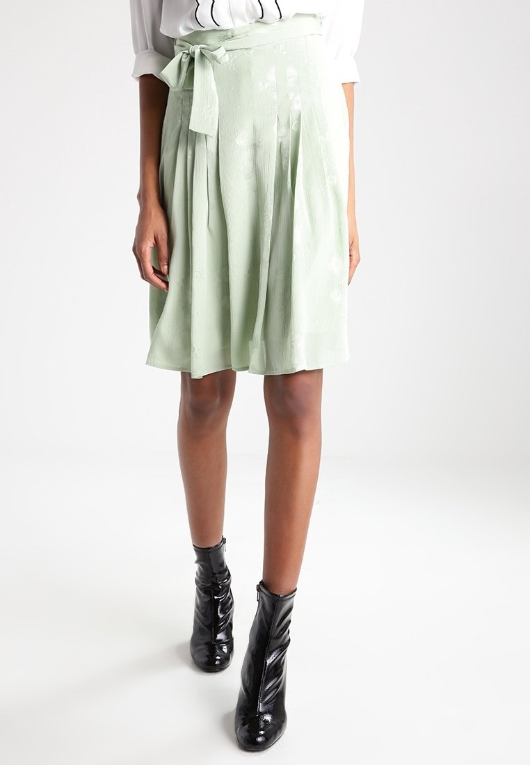 Femme portant une jupe vert clair arrivant au genou avec une ceinture nouée à la taille et des bottines noires, debout devant un fond blanc uni.