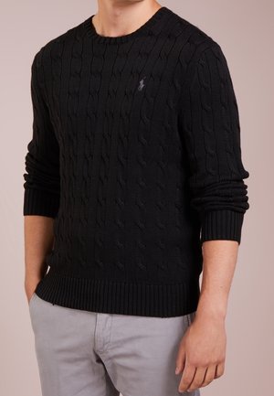 Man draagt zwarte kabelgebreide trui met klein geborduurd logo op de borst en lichtgrijze broek, handen ontspannen langs het lichaam.