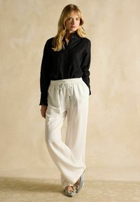 Blonde woman standing with hands in pockets wearing black buttoned blouse, white drawstring pants, and gray pointed shoes with buckles.