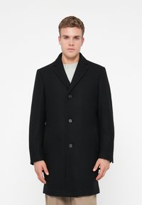 Un tânăr stă îmbrăcat cu un palton negru cu butoni peste o cămașă gri deschis și pantaloni bej, pe un fundal alb simplu.