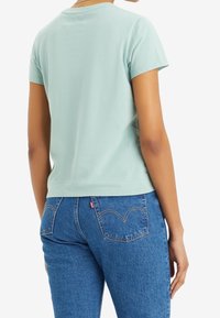 Svetle mátově zelené tričko s krátkým rukávem a kulatým výstřihem, spárované s modrými džínami s prošitými detaily na zadních kapsách.