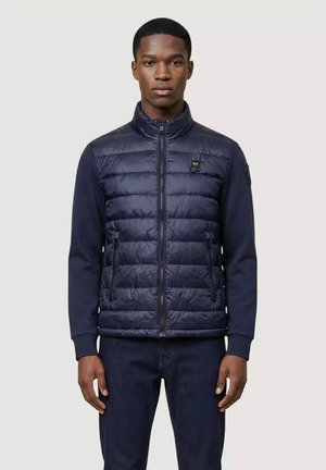 Giovane uomo che indossa una giacca trapuntata blu navy con zip frontale e maniche blu scuro, in piedi con le braccia rilassate lungo i fianchi.