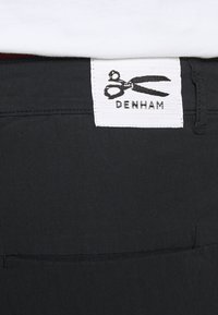 Tissu en denim noir avec une ceinture plate. Dispose d'une étiquette blanche avec "DENHAM" et un logo en forme de ciseaux. Couture simple, sans quincaillerie visible.
