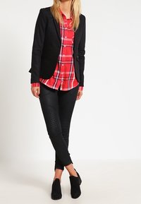 Svart kavaj över en röd rutmönstrad skjorta, ihop med åtsittande svarta byxor med kviltad textur, och svarta anklarstövlar.