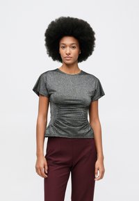 Žena s afro frizurom nosi metalik sivi top s kratkim rukavima i naborima, uz visoko strukirane bordo hlače, naslonjena na jednostavnu bijelu pozadinu.