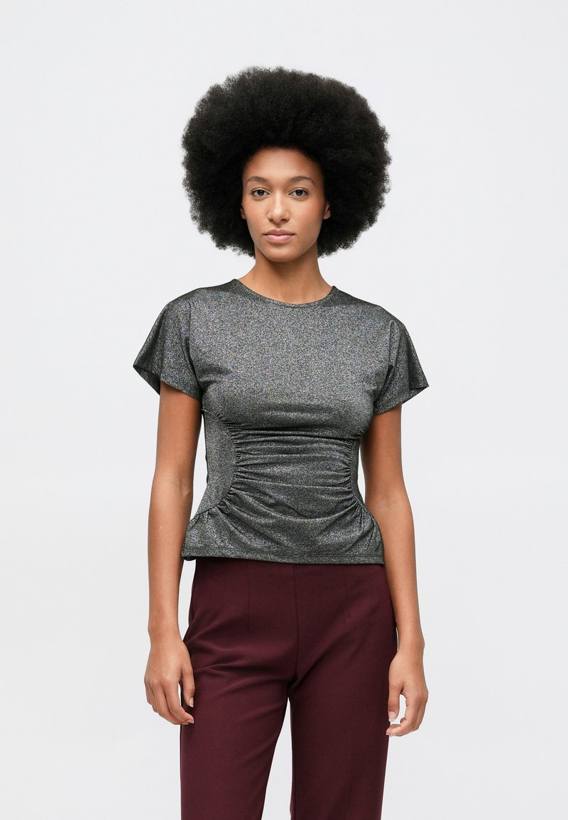 Žena s afro frizurom nosi metalik sivi top s kratkim rukavima i naborima, uz visoko strukirane bordo hlače, naslonjena na jednostavnu bijelu pozadinu.