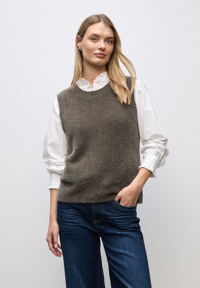 Grå, fluffig ärmlös tröja lager på lager över en vit skjorta med krage. Har rund halsringning och en avslappnad passform, med mörkblå jeans.