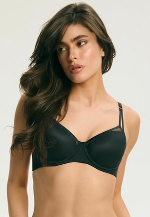 Kobieta o długich ciemnych włosach, ubrana w czarny biustonosz, patrząca w lewo na jasnym, jednolitym tle.