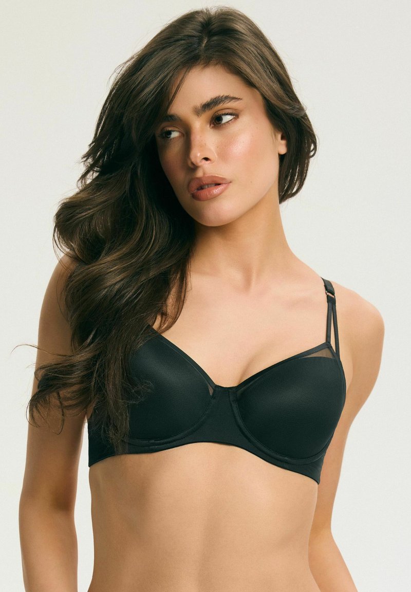 Žena s dlhými tmavými vlasmi, ktorá nosí čiernu podprsenku a pozerá sa doľava na jednoduchom svetlom pozadí.