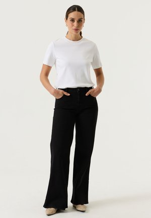Femme debout, les mains dans les poches, portant une chemise blanche à manches courtes, un pantalon noir à jambes larges, des chaussures beiges et des boucles d'oreilles créoles dorées.