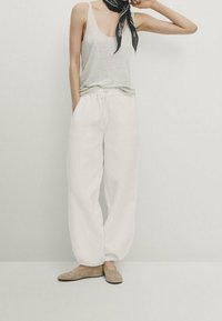 Witte, losse sweatpants met een elastische tailleband, gecombineerd met een lichtgrijze tanktop en zachte beige instapschoenen. Zwarte sjaal geknoopt om de nek.