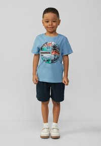 Jasnoniebieska bawełniana koszulka z okrągłym nadrukiem przedstawiającym palmy oraz napisem "SESJE ZACHODU SŁOŃCA". Połączona z ciemnymi szortami i białymi butami.