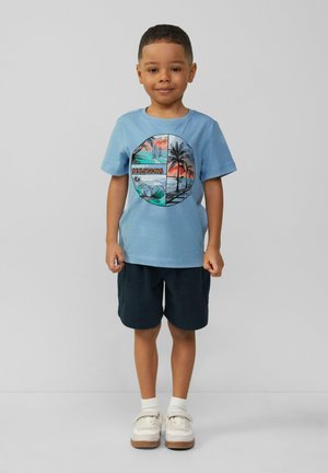 Lichtblauw katoenen T-shirt met een cirkelvormige afbeelding van palmbomen en de tekst "SUNSET SESSIONS." Gecombineerd met donkere shorts en witte schoenen.