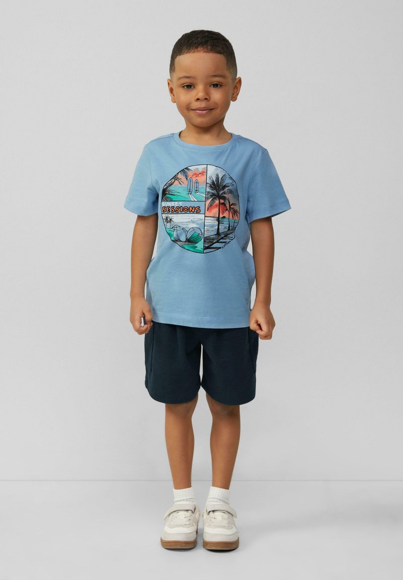 Jasnoniebieska bawełniana koszulka z okrągłym nadrukiem przedstawiającym palmy oraz napisem "SESJE ZACHODU SŁOŃCA". Połączona z ciemnymi szortami i białymi butami.