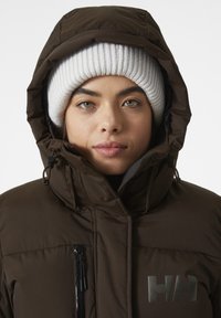Bruine gewatteerde jas met hoge kraag en capuchon, voorzien van een zwart ritsvak en een zilveren logo op de borst. De persoon draagt een witte geribbelde beanie.