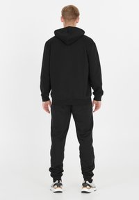 Mies seisoo selkä kameralle päin, päällä musta huppari ja verkkohousut, jalassaan valkoiset ja mustat tennarit valkoista taustaa vasten.