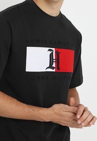 Camiseta de algodón negra con un logotipo destacado de las iniciales "H" en rojo y blanco, con el texto "LEWIS HAMILTON" y "TOMMY HILFIGER" debajo.
