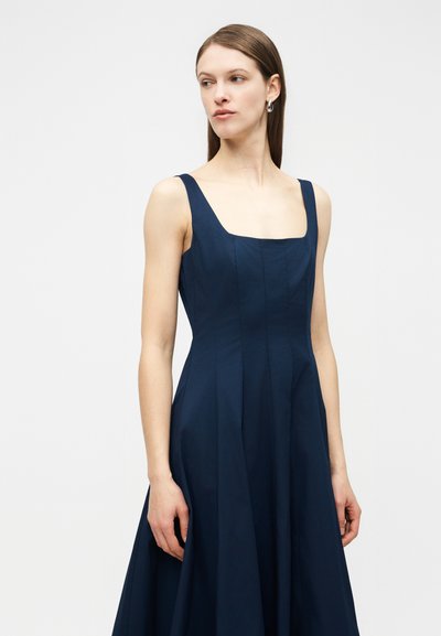 Γυναίκα που φοράει αμάνικο φόρεμα σε σκούρο μπλε με τετράγωνη λαιμόκοψη, στέκεται με τα χέρια χαλαρά, κοιτάζοντας ελαφρώς προς τα αριστερά της.