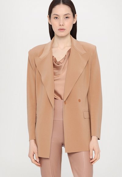 Beige kaksirivinen bleiseri leveillä kauluksilla, muotonsa mukaan räätälöity, yhdistettynä satiiniseen cowl-neck paitoihin ja samaan istuvaan housupariin. Tasainen pinta.