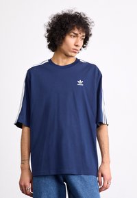 Marineblå oversized t-shirt lavet af bomuld med hvide tre-stribe detaljer på skuldrene. Har et lille hvidt Adidas-logo på brystet.