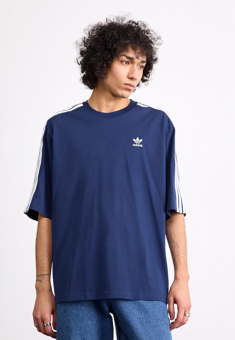 Marineblå oversized t-shirt lavet af bomuld med hvide tre-stribe detaljer på skuldrene. Har et lille hvidt Adidas-logo på brystet.
