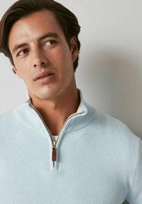 Šviesiai mėlynas užtrauktuku zipuojamas megztinis su gauromis apykakle ir odiniu akcentu prie užtrauktuko, pasižymintis megztu raštu ir prigludusia forma.